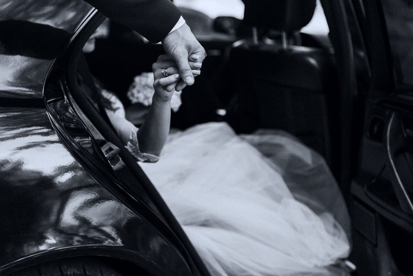 Detail of a bride's hand held by a groom at the open door of a black wedding car